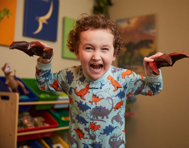 Dressed in gray pajamas covered with multi-colored dinosaurs, a 4-year-old child pretends to be a dinosaur roaring. In the background is a playroom, with a shelf full of toys and dinosaur images on the walls. 